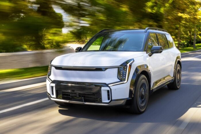 White Kia SUV driving on a scenic road with trees in the background.